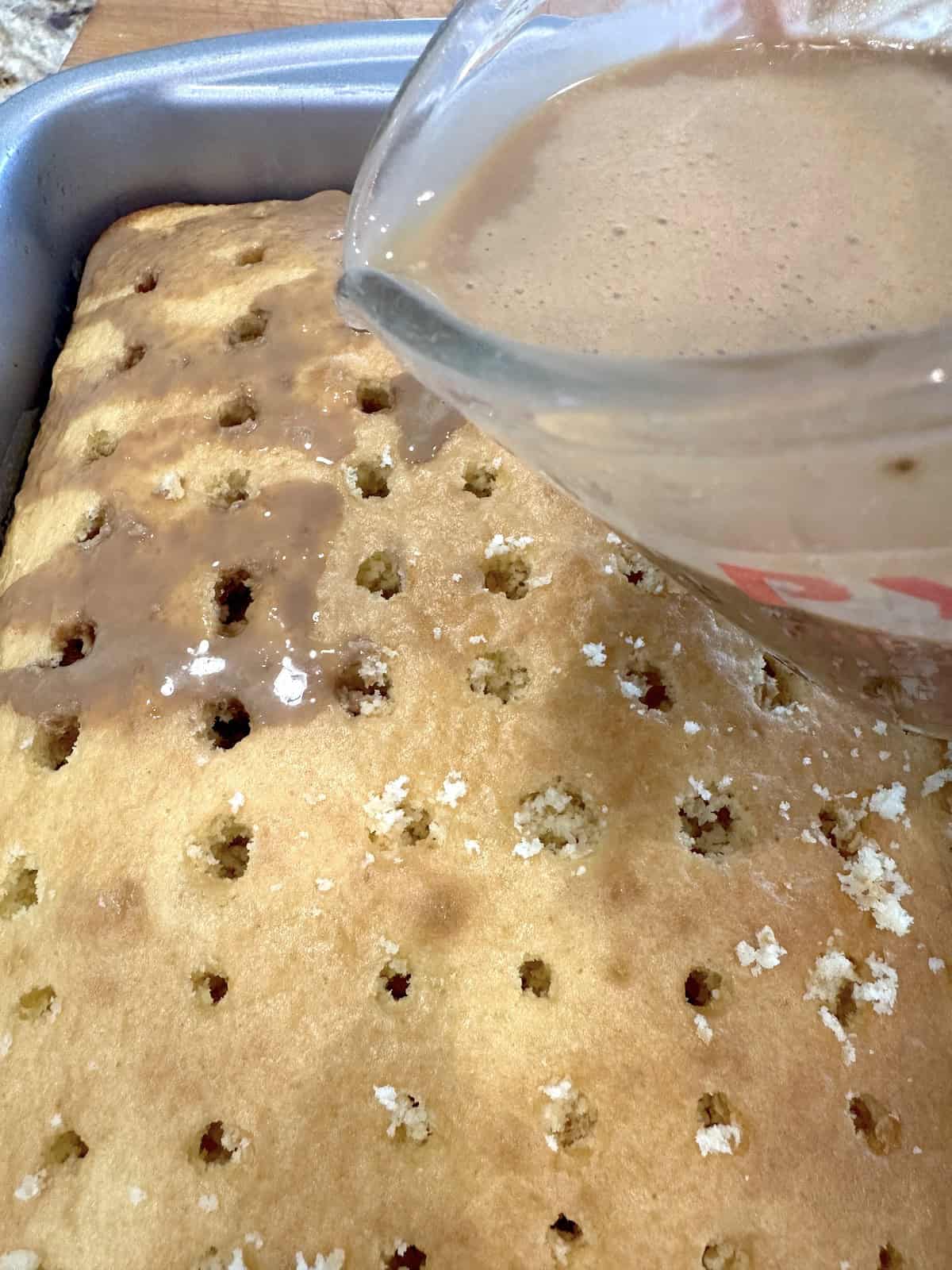 A measuring cup full of coffee mixture over a cake with holes.