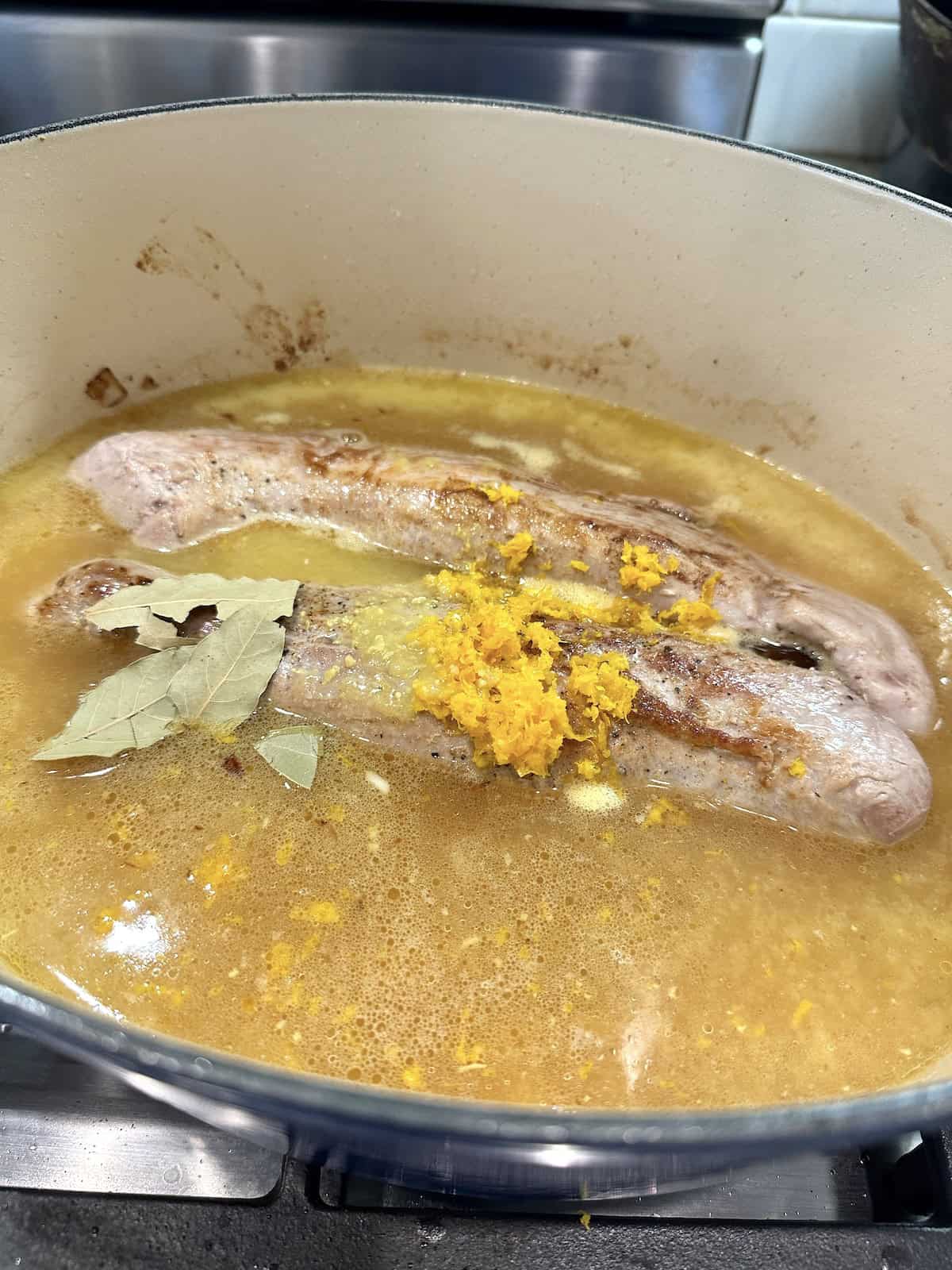 Whole pork tenderloins in a pan of orange sauce with orange zest and bay leaves.
