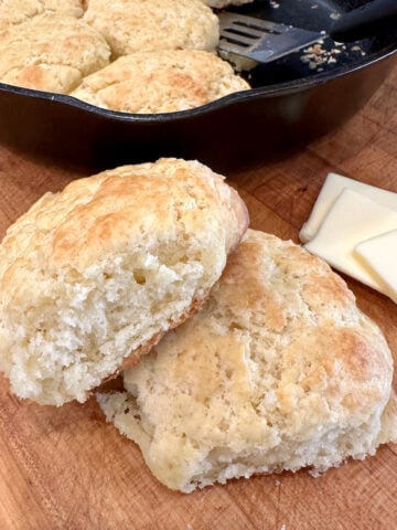 Baked homemade biscuits on a cutting board with a cast iron skillet and butter.